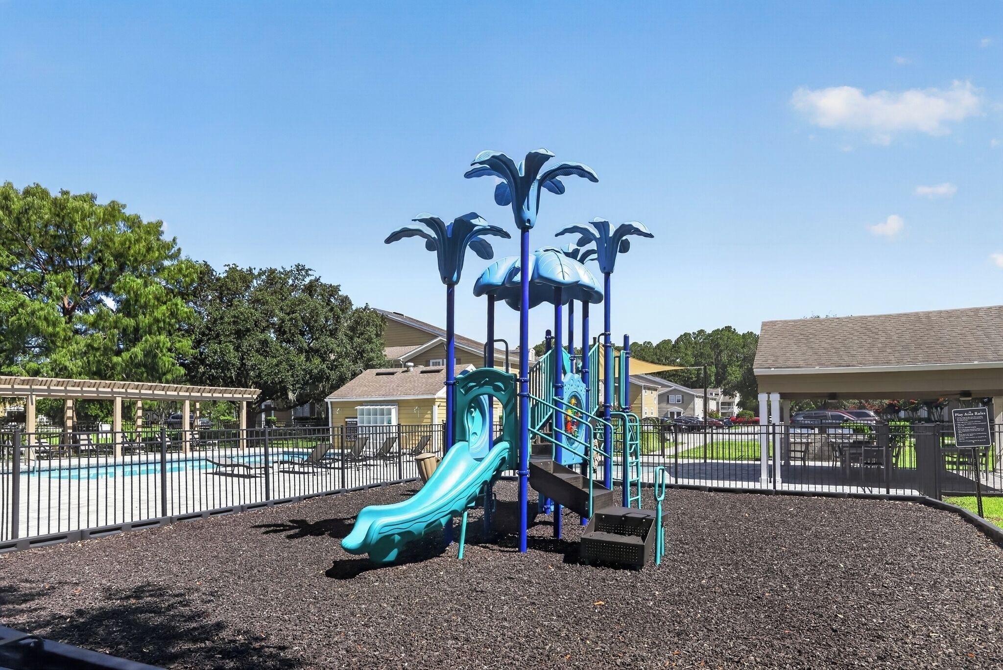 Un parque infantil con un tobogán y estructuras para escalar, rodeado por una valla y árboles.