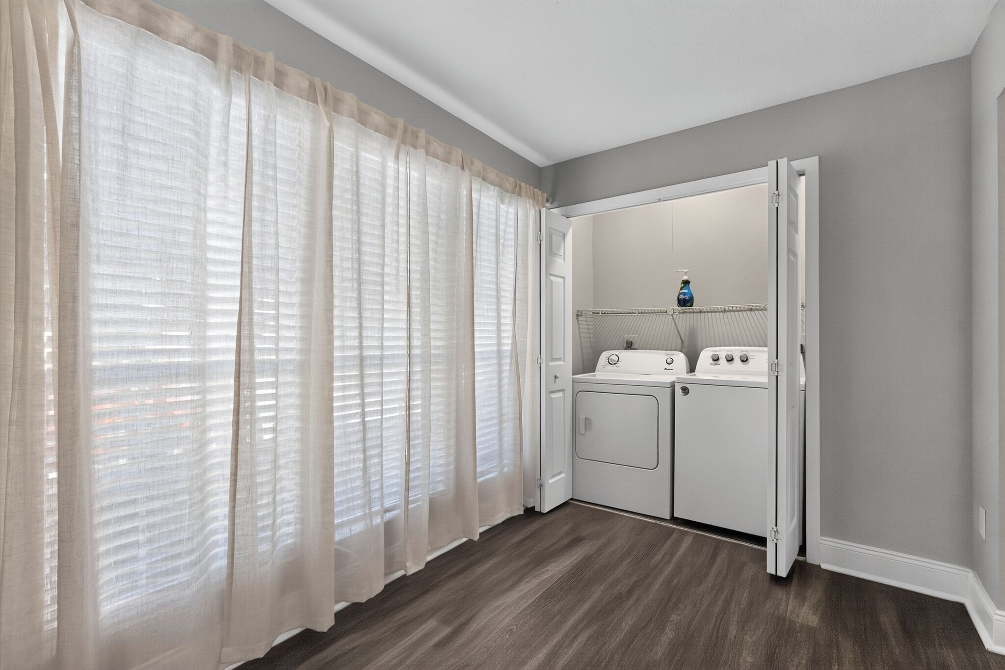 Una sala de lavandería con lavadora y secadora, una puerta y un ventanal con cortinas translúcidas.