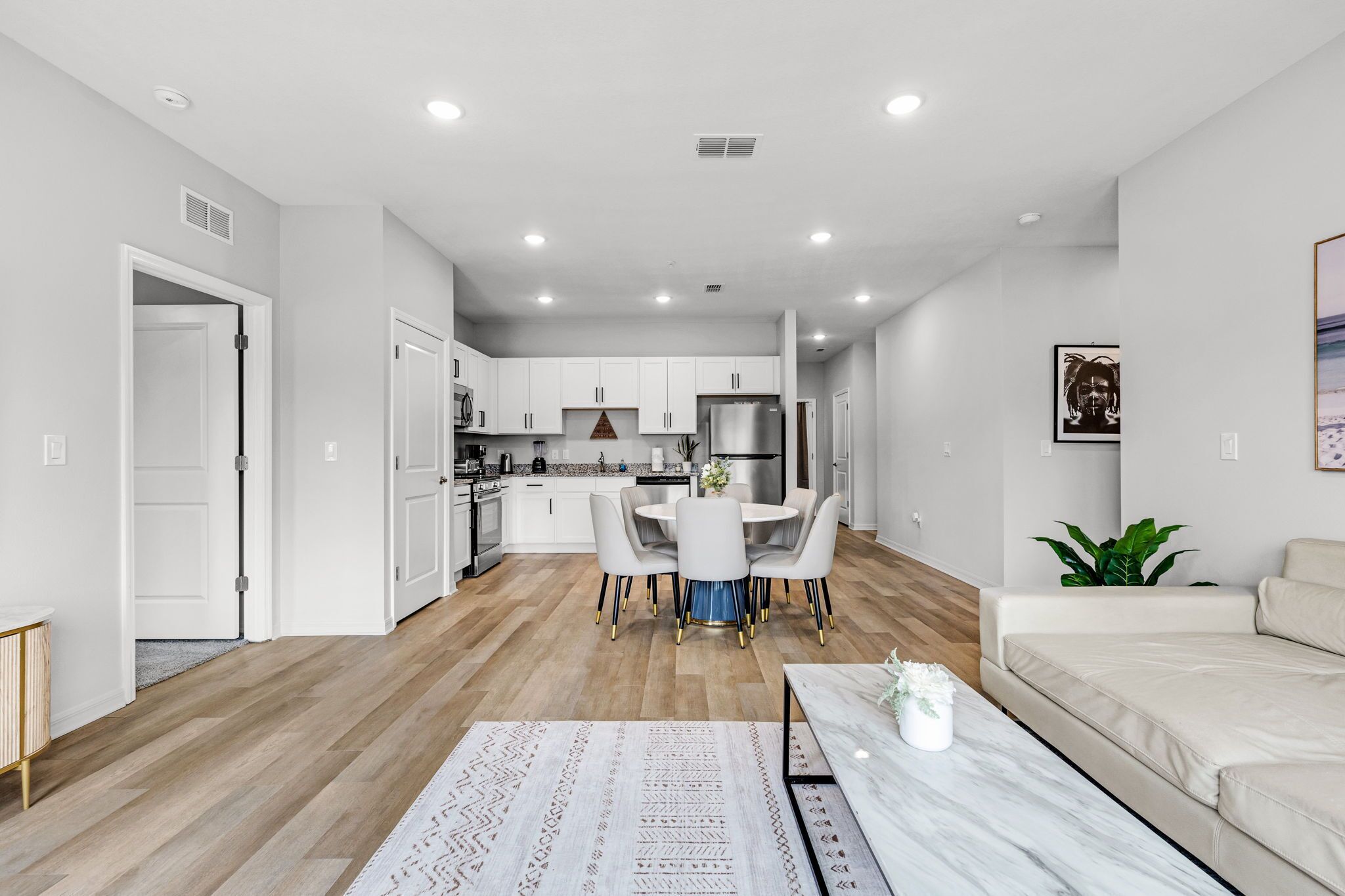 A modern kitchen with white cabinets, a dining area with a round table and chairs, a living room with a sofa, and a hallway leading to a door.