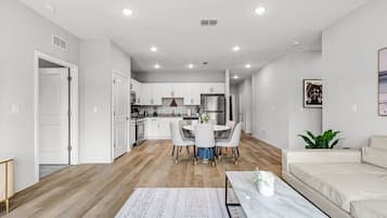 A modern kitchen with white cabinets, a dining area with a round table and chairs, a living room with a sofa, and a hallway leading to a door.