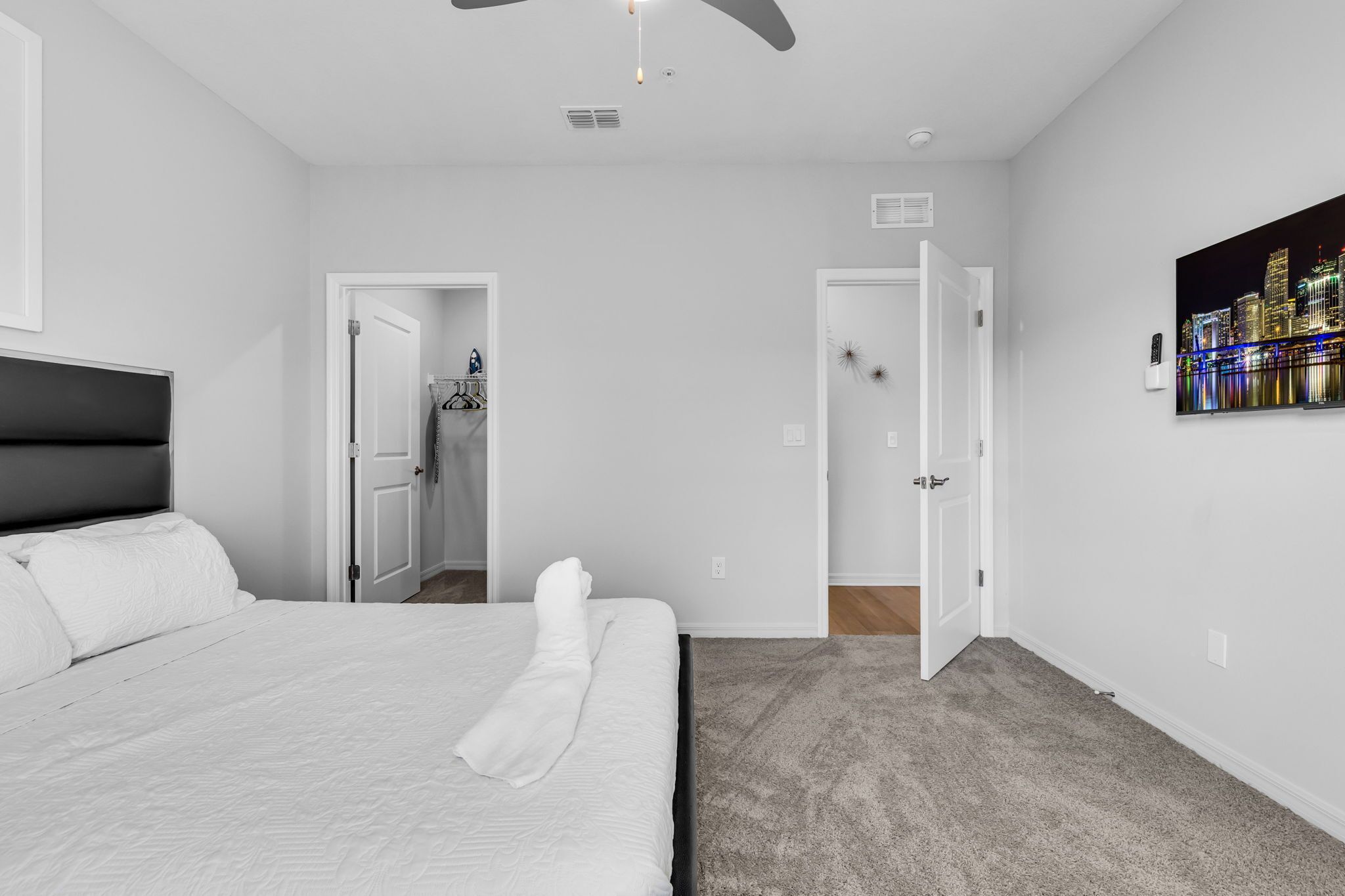 Een slaapkamer met een bed, een plafondventilator, een deur naar een badkamer en een stadsgezichtschilderij aan de muur.