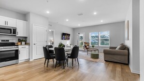A modern kitchen with stainless steel appliances, a dining area with black chairs, a grey sofa, and large windows.