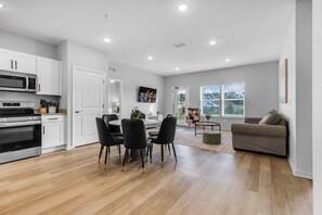 A modern kitchen with stainless steel appliances, a dining area with black chairs, a grey sofa, and large windows.