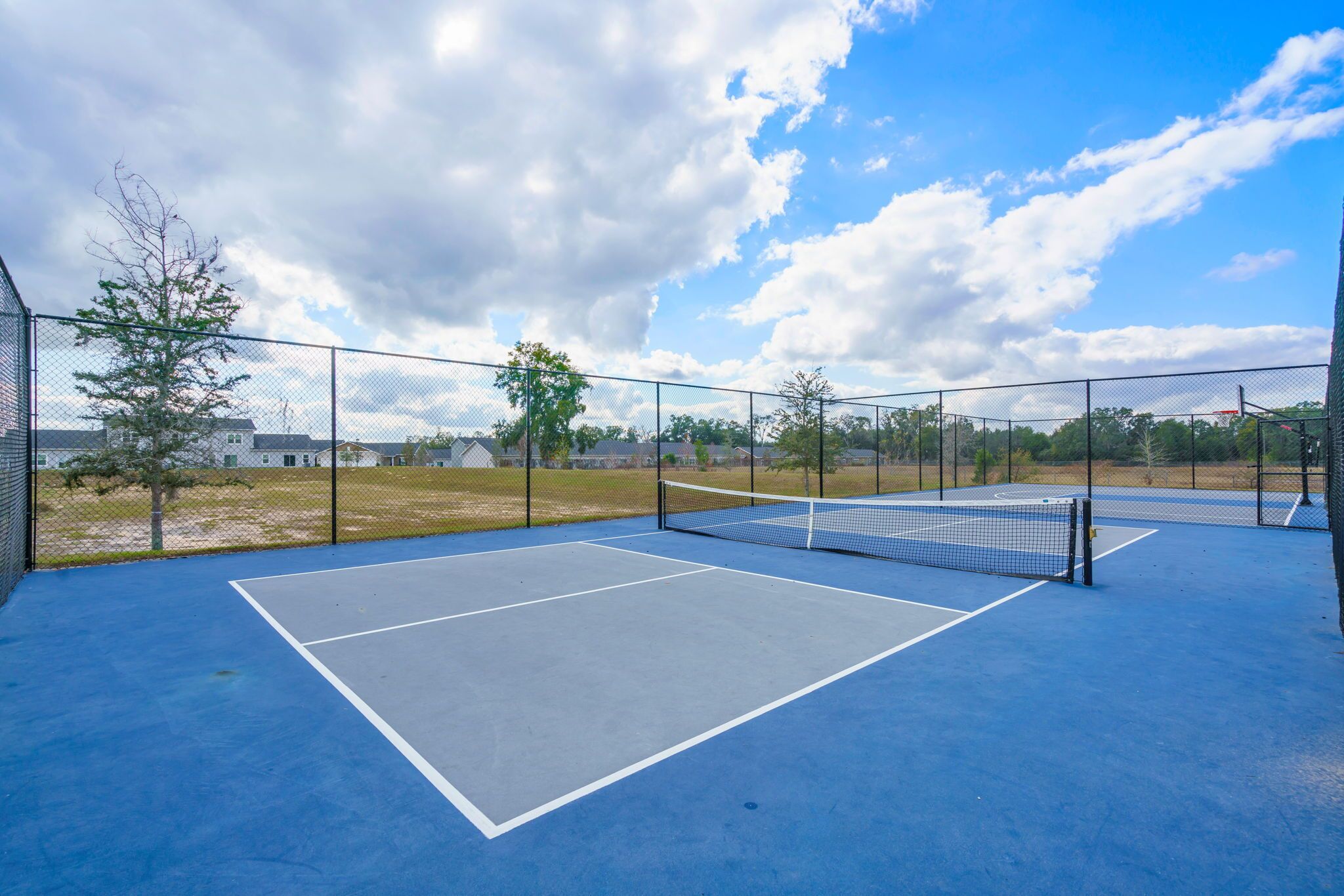 Una cancha de tenis al aire libre con una red, rodeada por una valla y árboles al fondo.