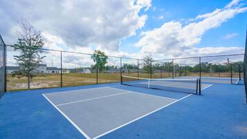 Una cancha de tenis al aire libre con una red, rodeada por una valla y árboles al fondo.