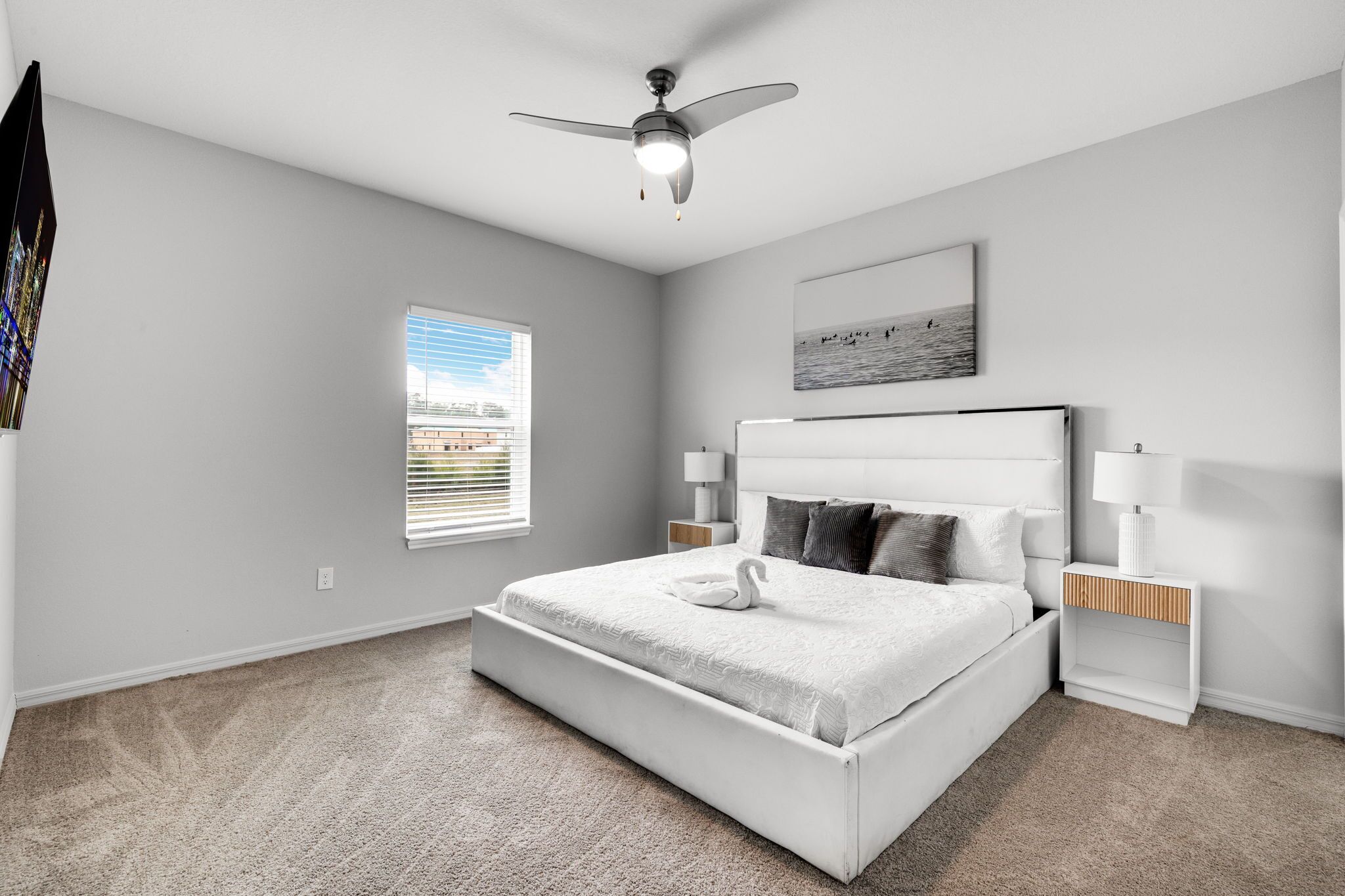 A bedroom with a bed, a ceiling fan, a window with blinds, a bedside table with a lamp, and a wall-mounted artwork.