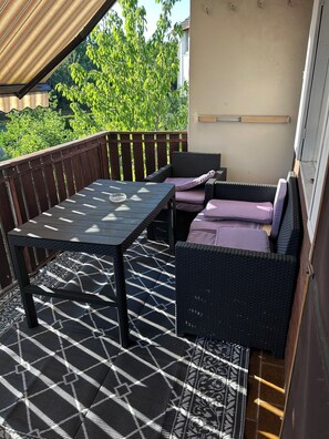 An outdoor patio with a table and wicker furniture, surrounded by a wooden deck and a view of greenery.