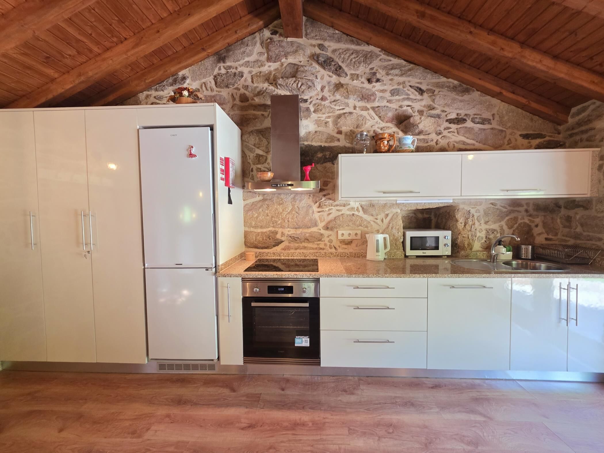 A kitchen with stone walls, wooden beams, a refrigerator, oven, microwave, and white cabinets.