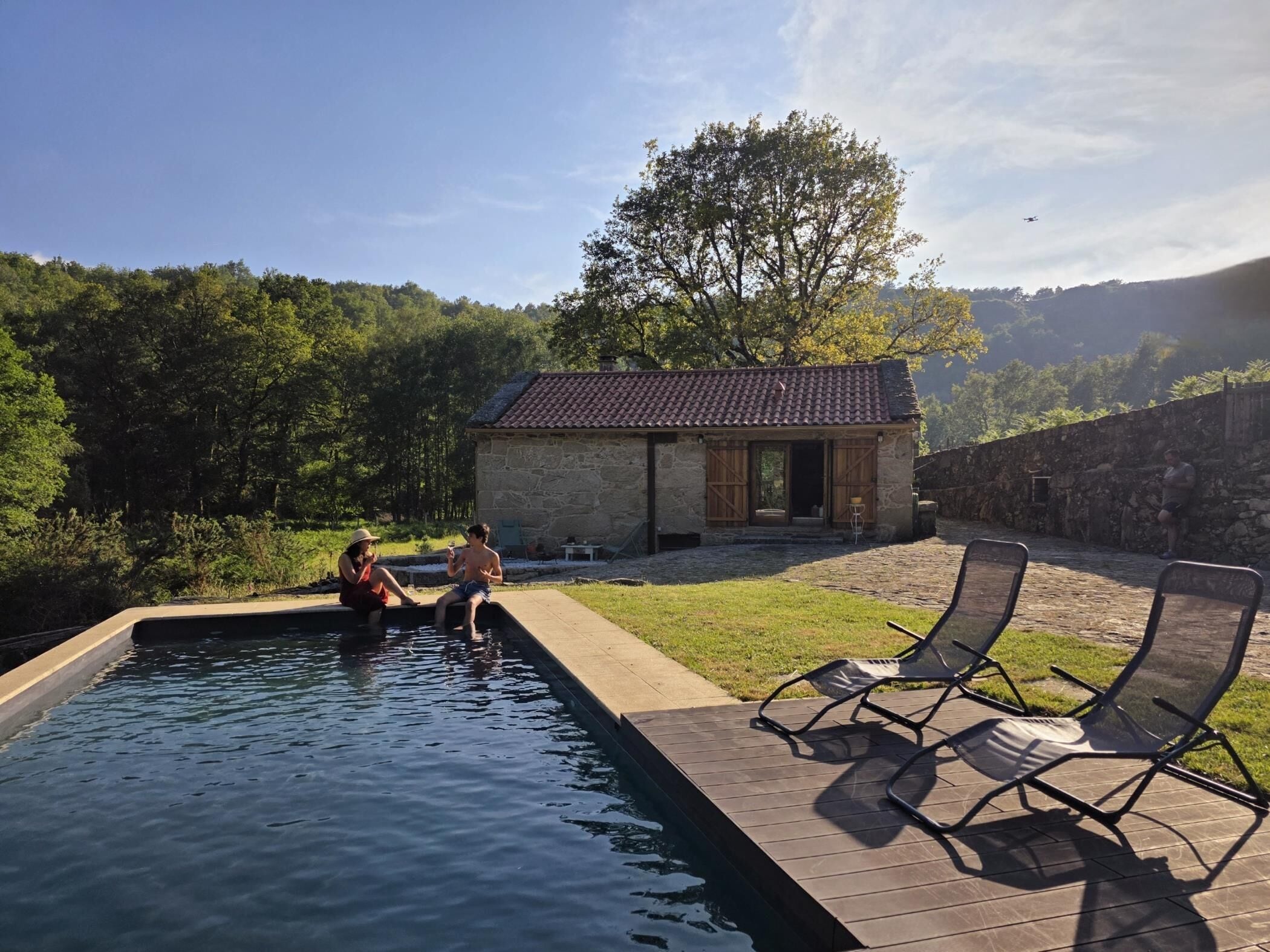 Una piscina con dos personas sentadas en el borde, un edificio de piedra y un árbol al fondo.