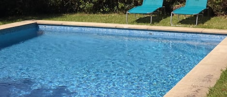 Une piscine rectangulaire à l’eau d’un bleu limpide, bordée d’une margelle en béton. Deux chaises longues vertes sont placées à l’extrémité de la piscine.