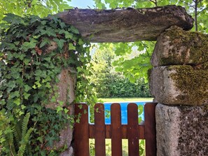 Ein hölzernes Tor mit einem blauen Schild, umgeben von Grünpflanzen und einer Steinstruktur.
