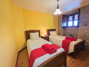 Ein Zimmer mit zwei Holzbettgestellen, einer Steinwand, einem Fenster mit Tageslicht und einer hängenden Leuchte.