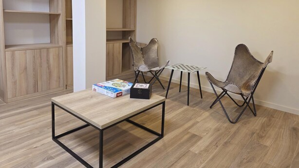A room with a wooden floor, a table with a box, two chairs, and a shelving unit. - Ponto Norte | Room 2 | Town Centre | Melgaço | The Northernmost Town in Portugal (Melgaço)