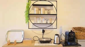 Una cocina con cafetera, tetera, bandeja a cuadros y estante con frascos y utensilios.