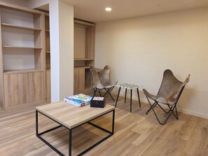 Una stanza con pavimento in legno, un tavolo con una scatola e un oggetto nero, due sedie e una libreria.