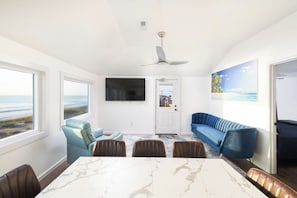 Ein modernes Wohnzimmer mit großem Fenster, Deckenventilator, Flachbildfernseher, blauem Sofa und Essbereich mit Marmortisch.