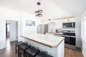 A modern kitchen with a marble island, stainless steel appliances, and a view into the adjacent living area. - Atlantic Inspiration @ Kitty Hawk Ocean Front (Kitty Hawk)