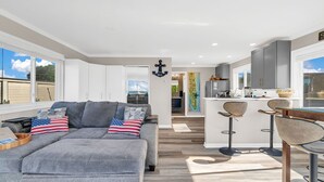 Uma sala de estar moderna com um sofá cinza, almofadas com a bandeira americana, uma mesa de centro de madeira e uma cozinha com armários cinzas e um banquinho.