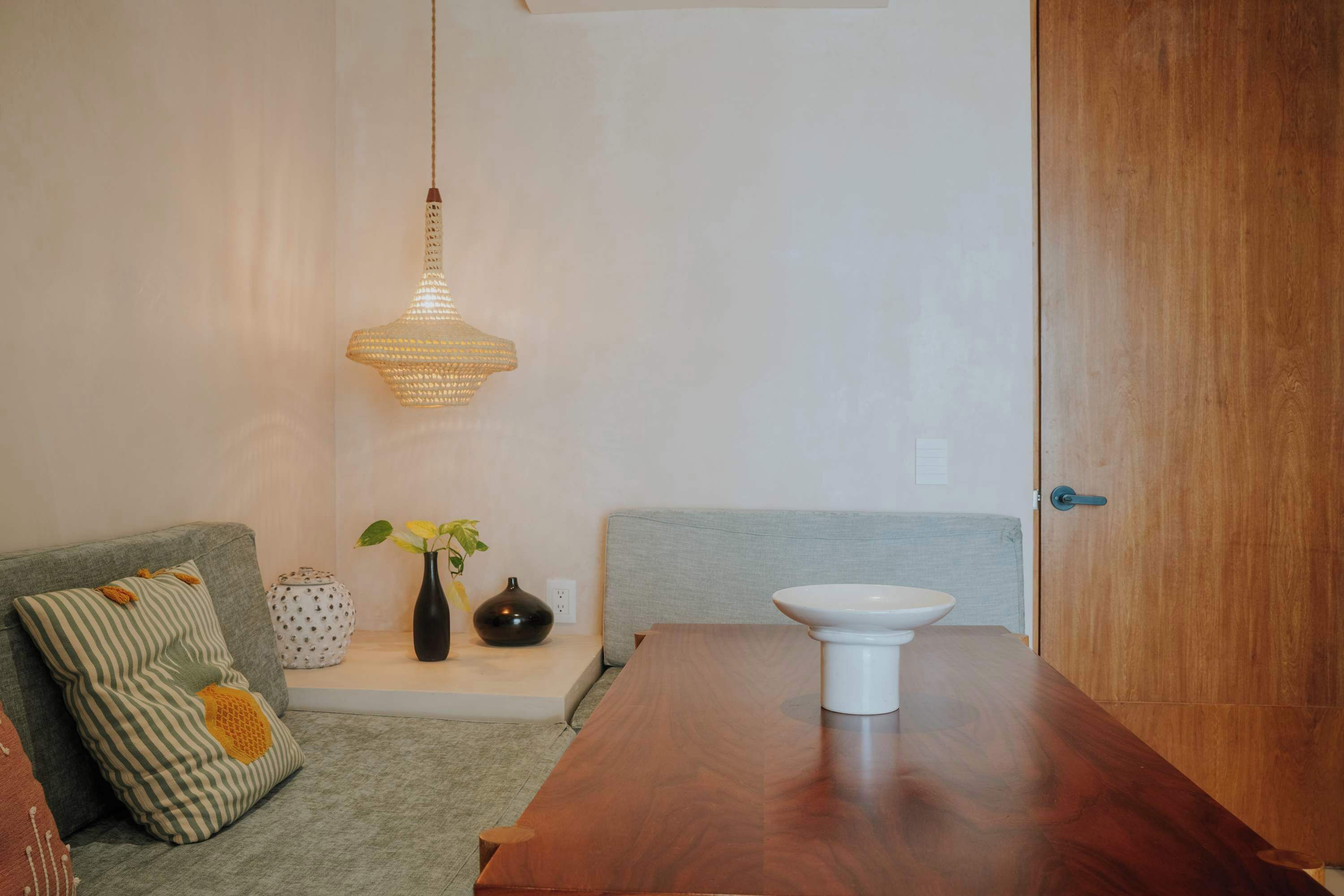 Een eethoek met een houten tafel, een grijze bank met gestreepte kussens, een hanglamp en een vaas met bloemen.