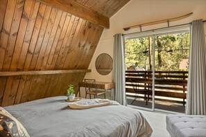 Ein Schlafzimmer mit schrägen Wänden, einem Bett, einem kleinen Tisch mit Tablett, einem Stuhl und einem Balkon mit Blick auf Bäume.