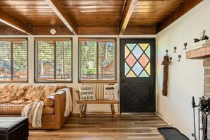 L’intérieur d’une cabane avec un plafond en bois, un canapé, un banc, une porte ornée d’un vitrail et un mur sur lequel sont accrochés divers objets.