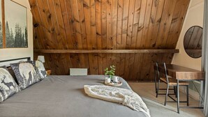 Un dormitorio con techo inclinado, una cama con almohadas estampadas, una mesita de noche con una planta y una silla.