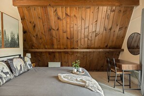 A bedroom with a sloped ceiling, a bed with patterned pillows, a bedside table with a plant, and a chair. - Solheim Lodge| Hot Tub, Fire Pit, EV Charger, Pets (Big Bear Lake)