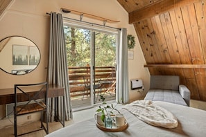 A cozy room with a bed, a chair, a table with a tray, a mirror, and a sliding glass door leading to an outdoor deck. - Solheim Lodge| Hot Tub, Fire Pit, EV Charger, Pets (Big Bear Lake)