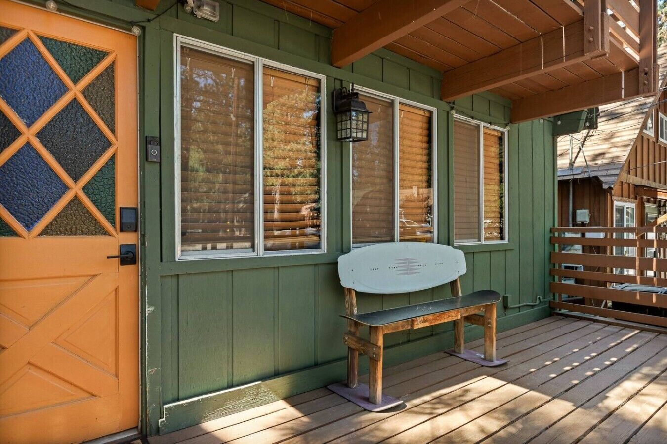 Una terraza de madera con un banco, una puerta con un panel de vidrio con diseño de rombos y un edificio verde con ventanas grandes.