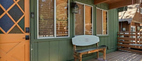 Una terraza de madera con un banco, una puerta con un panel de vidrio con diseño de rombos y un edificio verde con ventanas grandes.