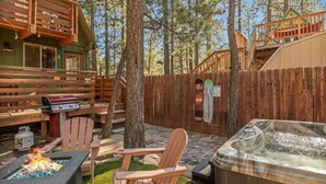 Terraza de madera con una parrilla, jacuzzi y zona de estar rodeada de árboles.