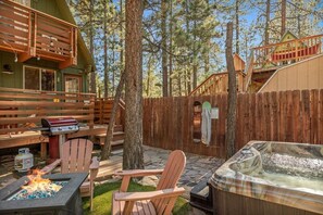 A wooden deck with a grill, hot tub, and seating area surrounded by trees. - Solheim Lodge| Hot Tub, Fire Pit, EV Charger, Pets (Big Bear Lake)