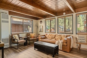 A cabin interior with wooden floors, a leather sofa, a wooden table, and large windows with blinds. - Solheim Lodge| Hot Tub, Fire Pit, EV Charger, Pets (Big Bear Lake)