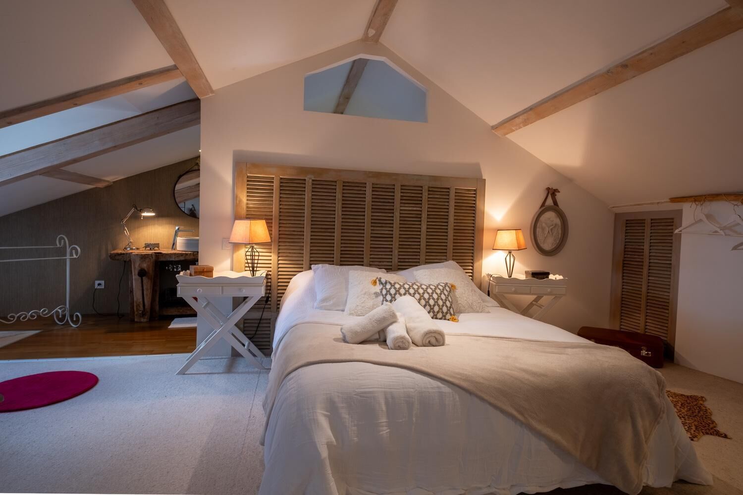 A bedroom with a sloped ceiling, a bed with white bedding, two bedside lamps, a small table, and a red yoga mat.