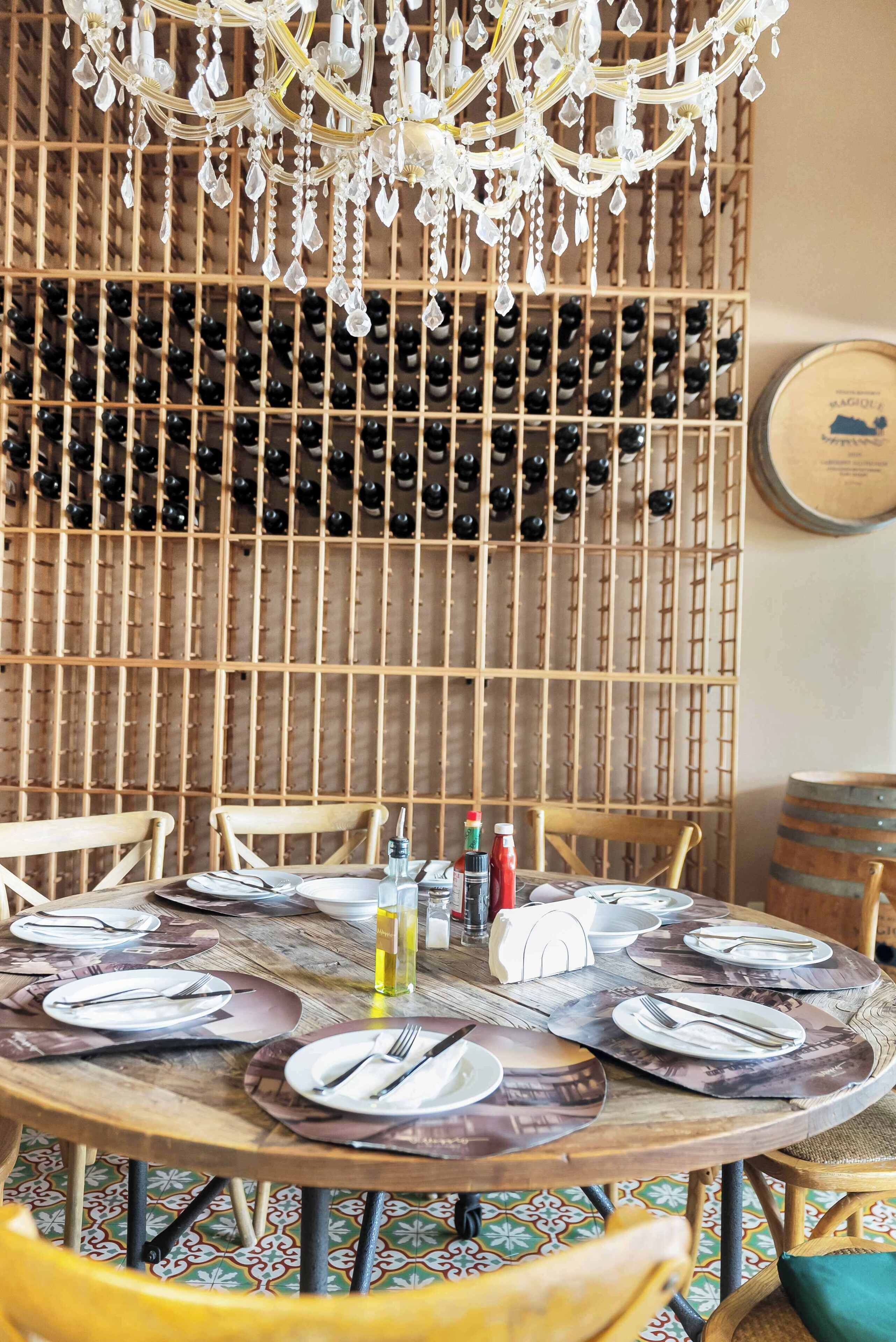 A dining area with a round table set for six, surrounded by chairs, and a wine rack in the background.