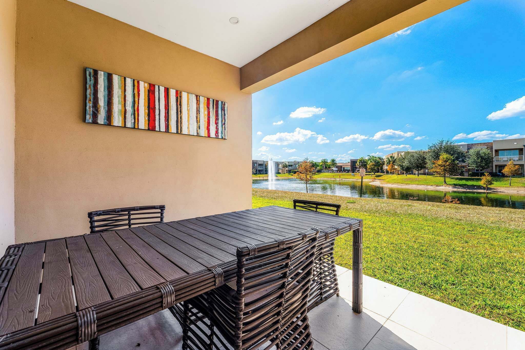 A patio with a table and chairs, a colorful abstract painting on the wall, and a view of a building complex and water feature.