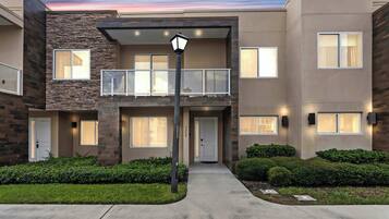 A modern multi-story residential building with a balcony, a front door, and a well-maintained lawn.