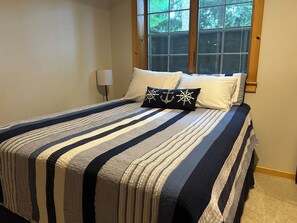 A bed with striped bedding, a window with a view of trees, and a bedside lamp. - Leland Cottage Walkable to Beach & Town (Leland)