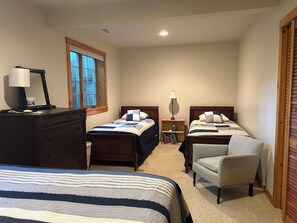 A bedroom with two beds, a dresser, a chair, a nightstand with a lamp, and a window with blinds.