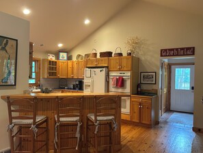 A kitchen with wooden cabinets, a central island with bar stools, a refrigerator, and a sign on the wall. - Leland Cottage Walkable to Beach & Town (Leland)