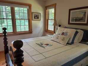 Ein Schlafzimmer mit hölzernem Bettgestell, einer gesteppten Bettdecke und zwei Fenstern mit Blick auf Grünflächen.