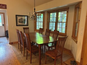 A dining room with a large wooden table surrounded by chairs, a chandelier, and large windows with a view of trees.