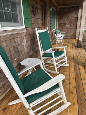 A wooden deck with white rocking chairs and green cushions, a small table, and a bear statue. - Leland Cottage Walkable to Beach & Town (Leland)