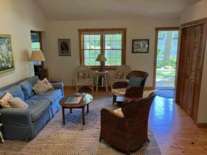 A living room with a sofa, chairs, a coffee table, and a rug. There are windows and a door leading outside.