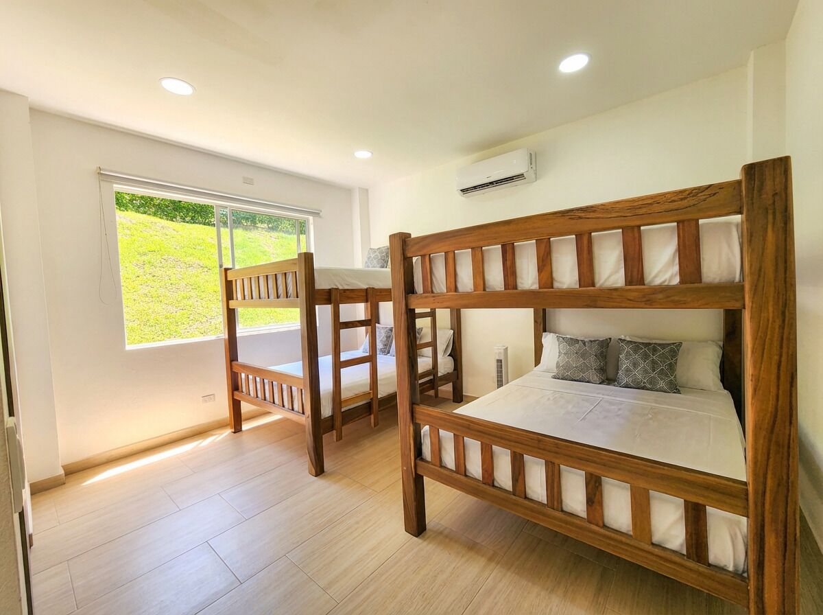 A room with two wooden bunk beds, a window with a view of greenery, and an air conditioning unit on the wall.