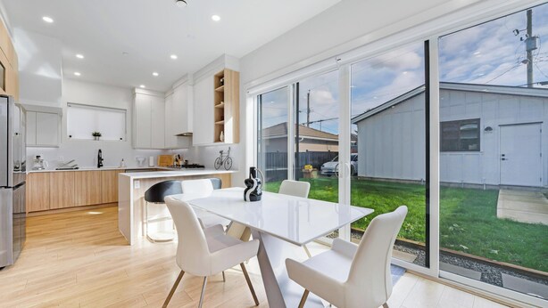 A modern kitchen with white cabinets, a dining area with a white table and chairs, and large windows offering a view of the backyard. - Modern 3BR Gem | Close to Transit | 15m to DT (Vancouver)