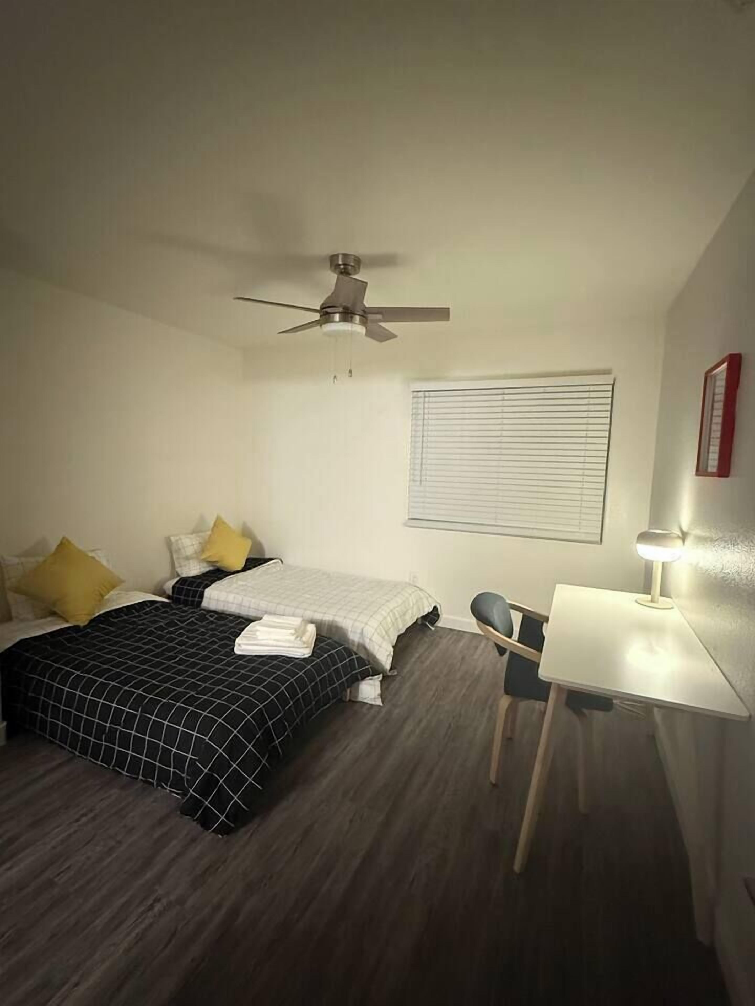 A bedroom with two beds, a ceiling fan, a desk with a chair, and a window with blinds.
