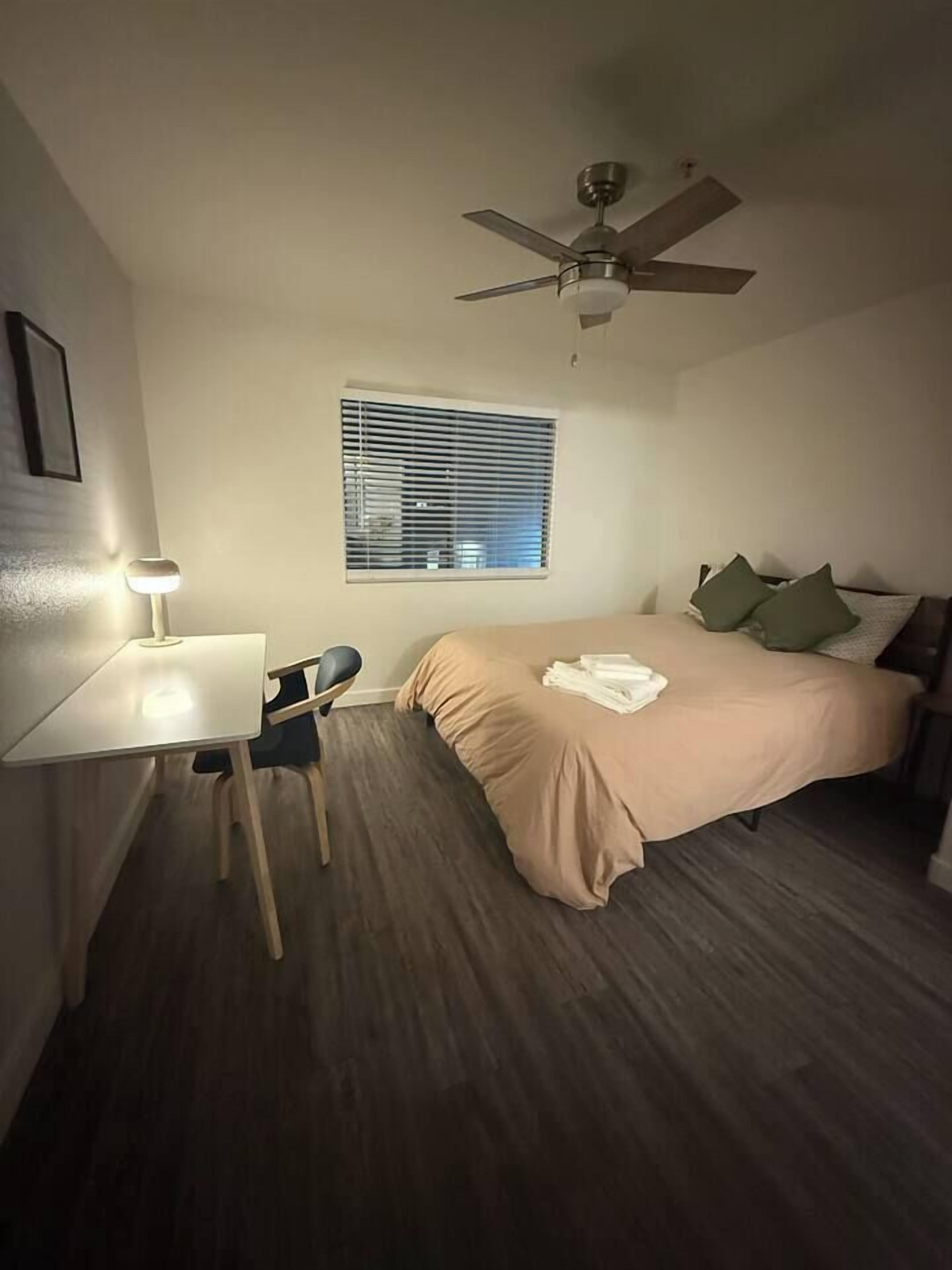 A bedroom with a bed, a desk with a chair, a ceiling fan, and a window with blinds.