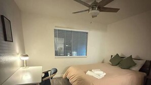 Un dormitorio con una cama, un escritorio con silla, un ventilador de techo y una ventana con persianas.