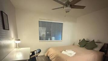 A bedroom with a bed, a desk with a chair, a ceiling fan, and a window with blinds.